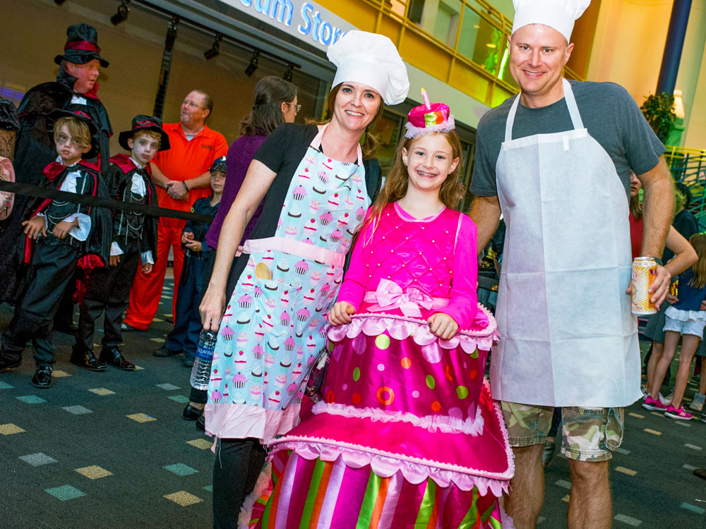 Child dressed in a pink costume that looks like she's a giant cake. Two grown-ups beside her are wearing chef costumes.